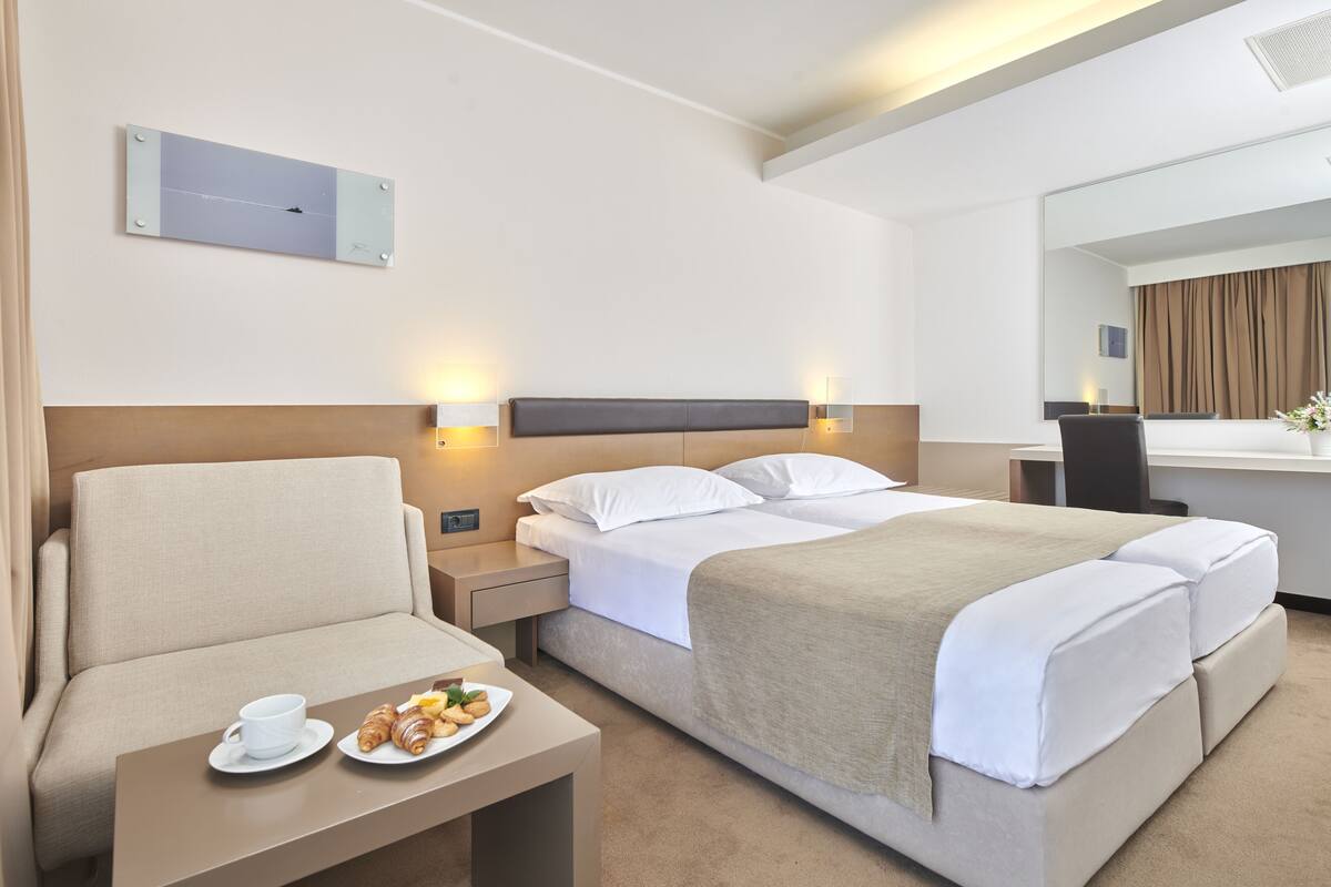 Quarto de hotel com cama, sofá, mesa pequena com xícara e doces, e espelho.