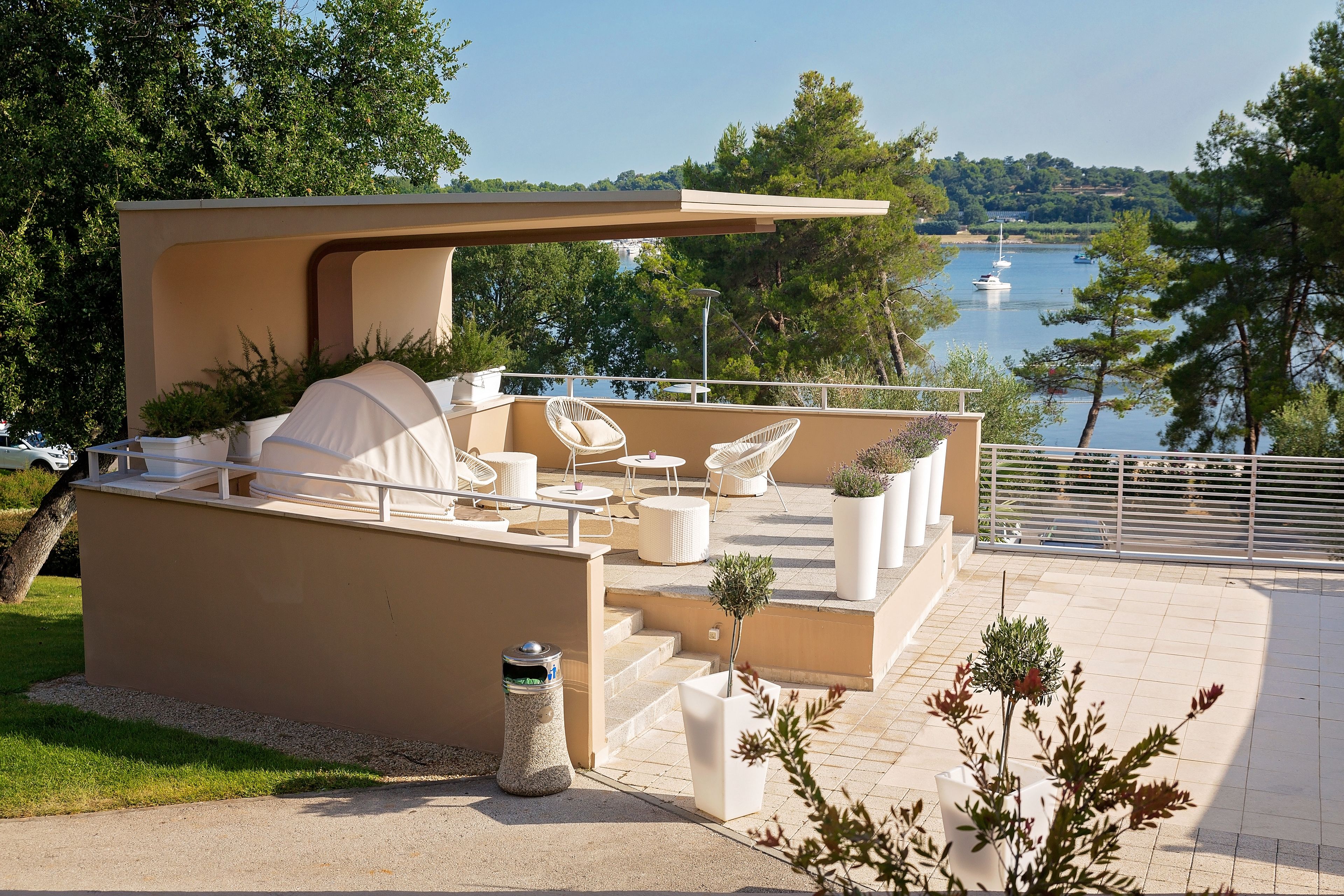 Eine moderne Terrasse im Freien mit Seeblick, ausgestattet mit weißen Stühlen und Tischen und von viel Grün umgeben.