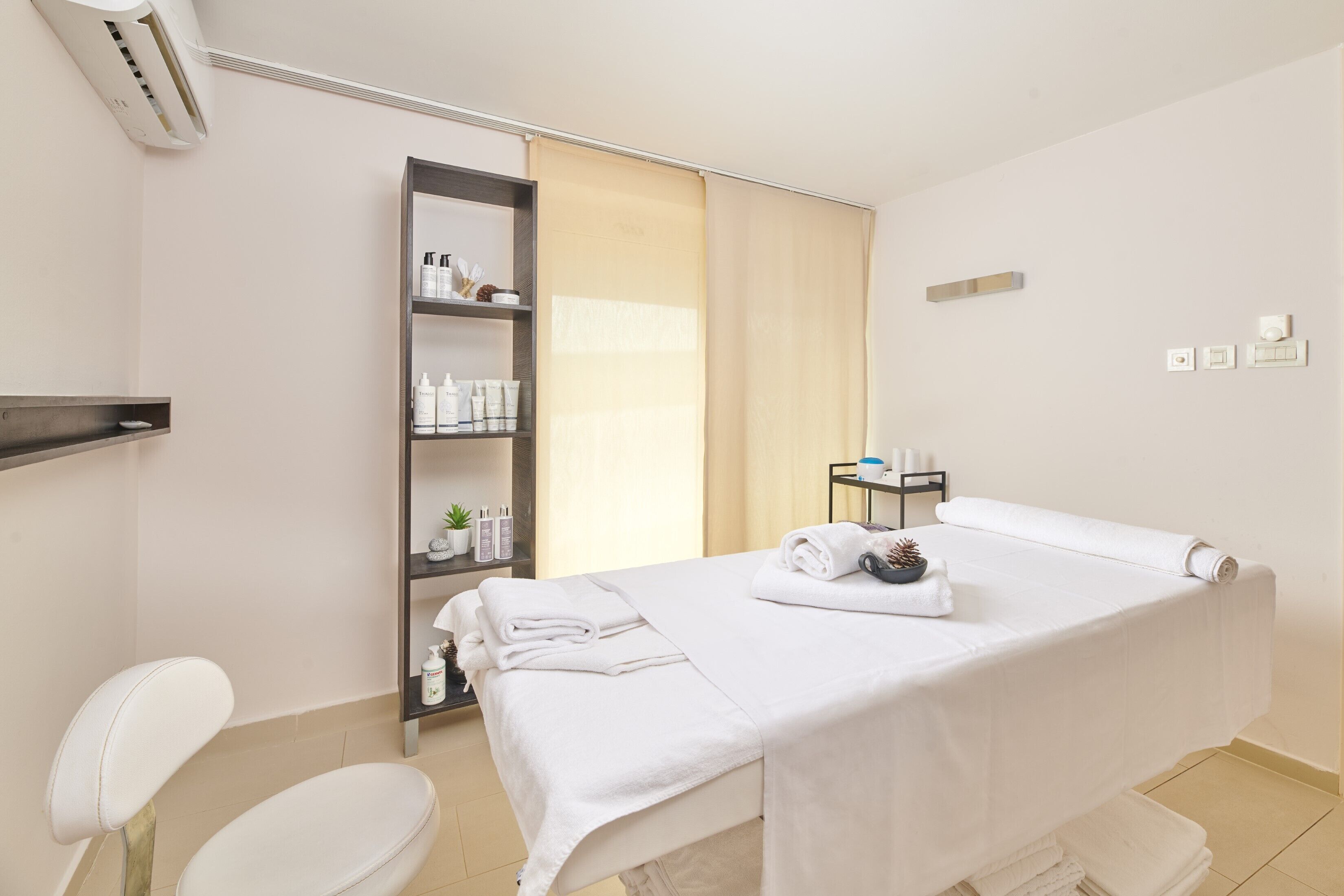 A clean, modern room with a massage table, a shelf with toiletries, and a window with curtains.