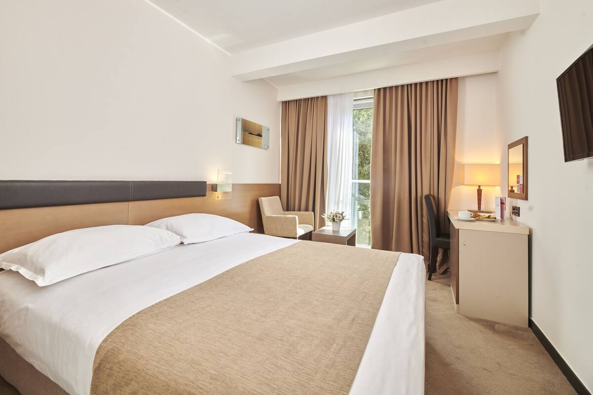 Quarto de hotel com uma cama grande, uma escrivaninha, uma cadeira e uma janela com cortinas.