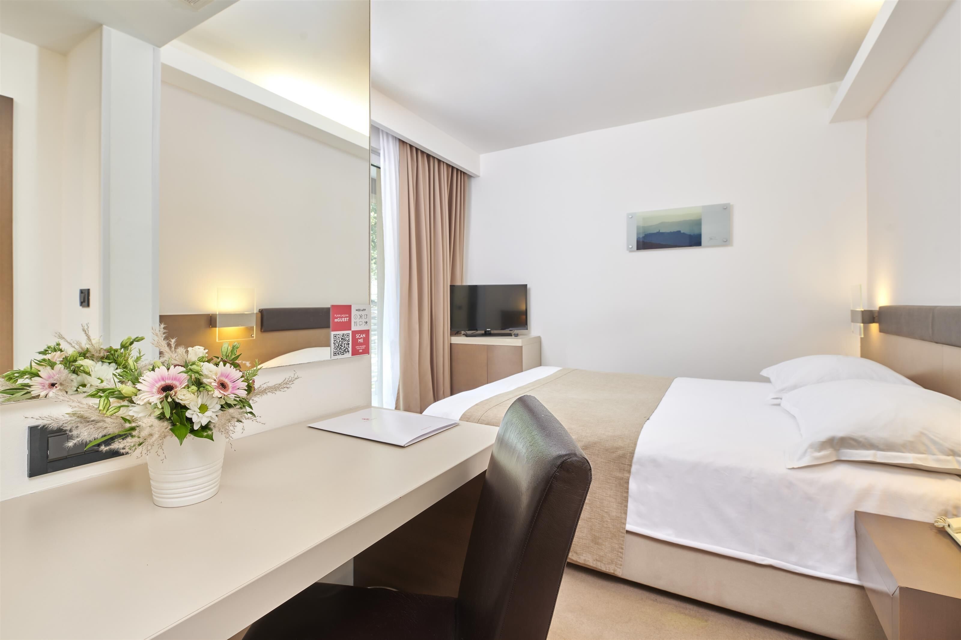 Habitación de hotel con cama, escritorio, laptop, televisor y un arreglo floral.