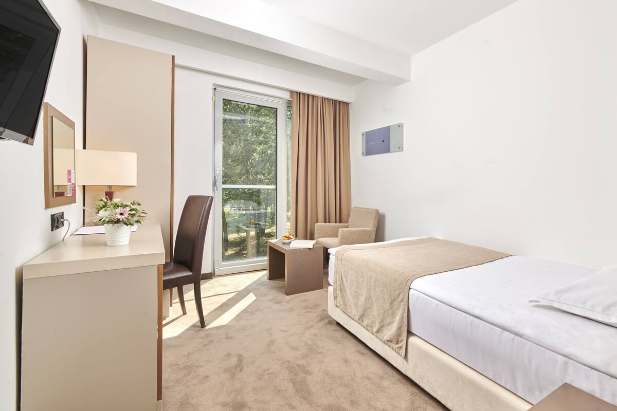 Quarto de hotel com cama, escrivaninha com luminária, cadeira, mesinha e porta de vidro deslizante que dá para uma varanda.
