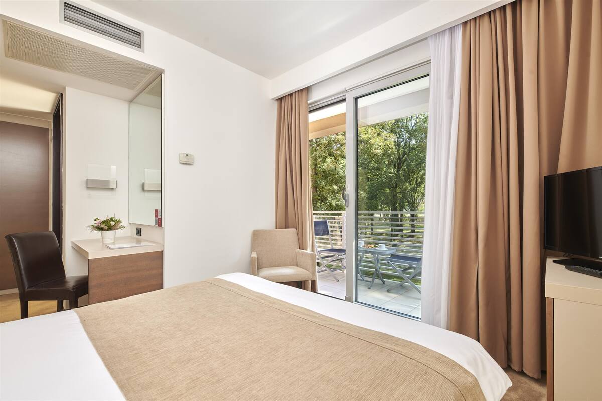 Quarto de hotel com cama, escrivaninha, cadeira, televisão e porta de vidro deslizante para área externa.