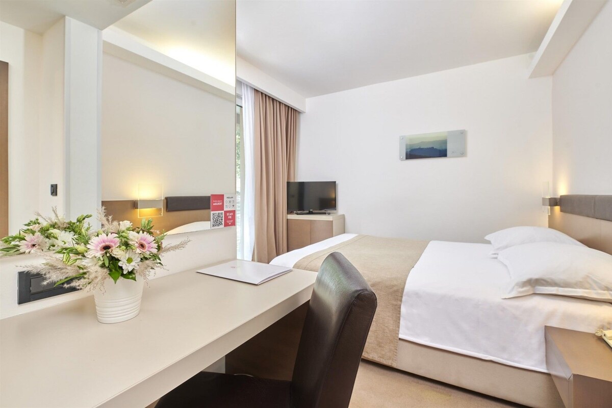 Una habitación de hotel con cama, escritorio con silla, televisor y ventana con cortinas.