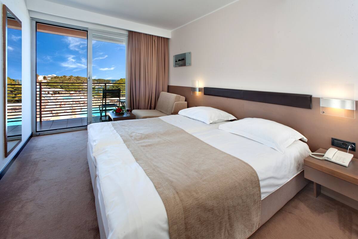 Quarto de hotel com uma cama grande, mesa de cabeceira com telefone, luz acoplada à parede e vista para uma piscina e vegetação através de uma porta de correr.