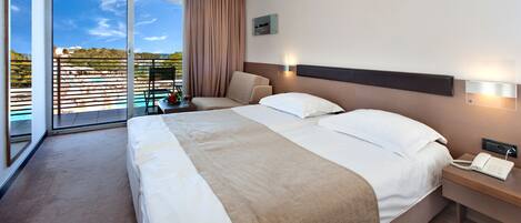 A hotel room with a large bed, a bedside table with a phone, a view of a pool and greenery through the window.
