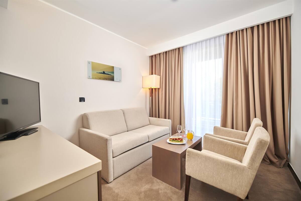 Quarto de hotel com televisão de tela plana, um sofá bege, uma mesa pequena com um prato de comida e um copo de suco, uma luminária e uma janela com cortinas.