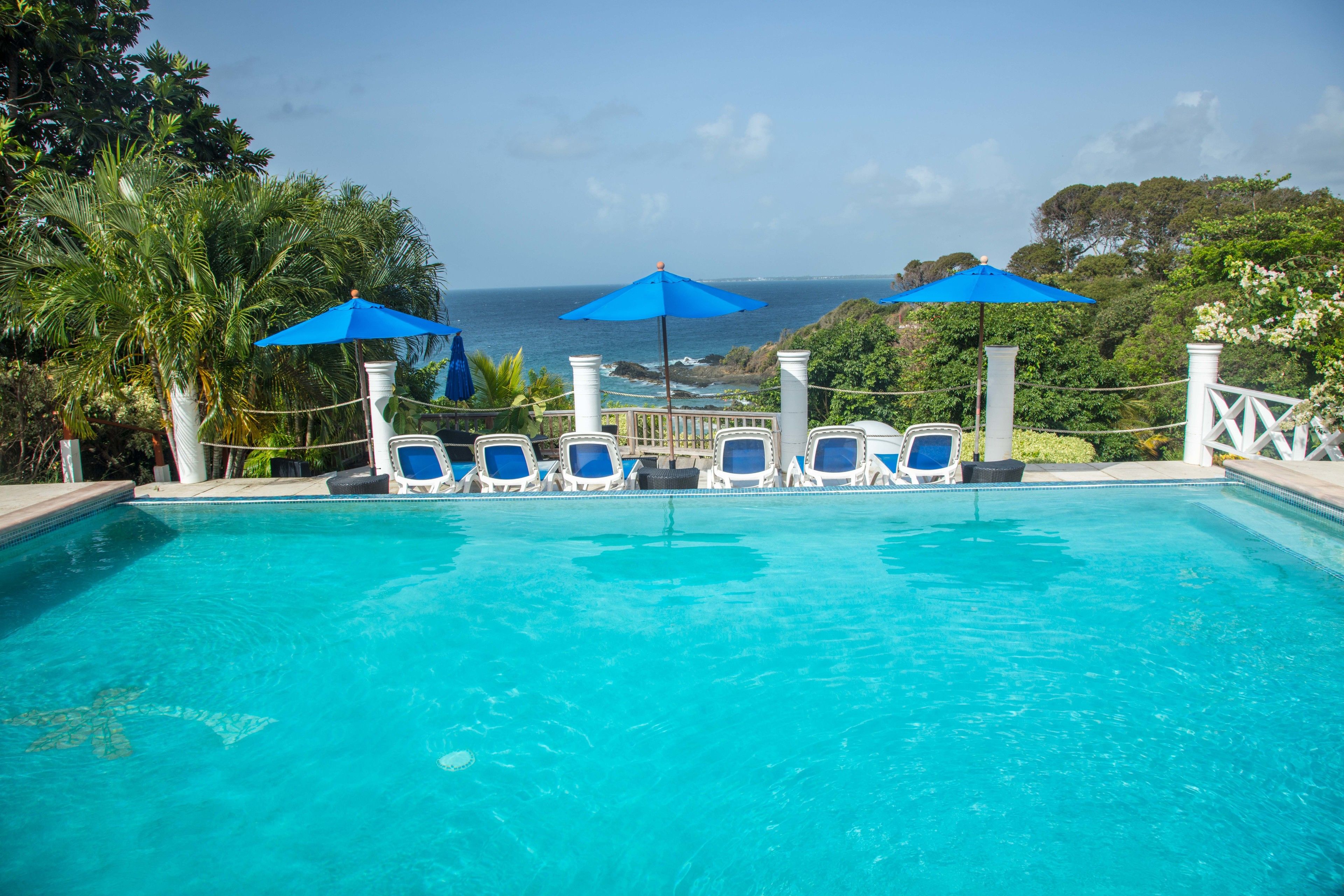 Ein Pool mit blauen Sonnenschirmen und Liegestühlen, umgeben von viel Grün und mit Blick auf das Meer.