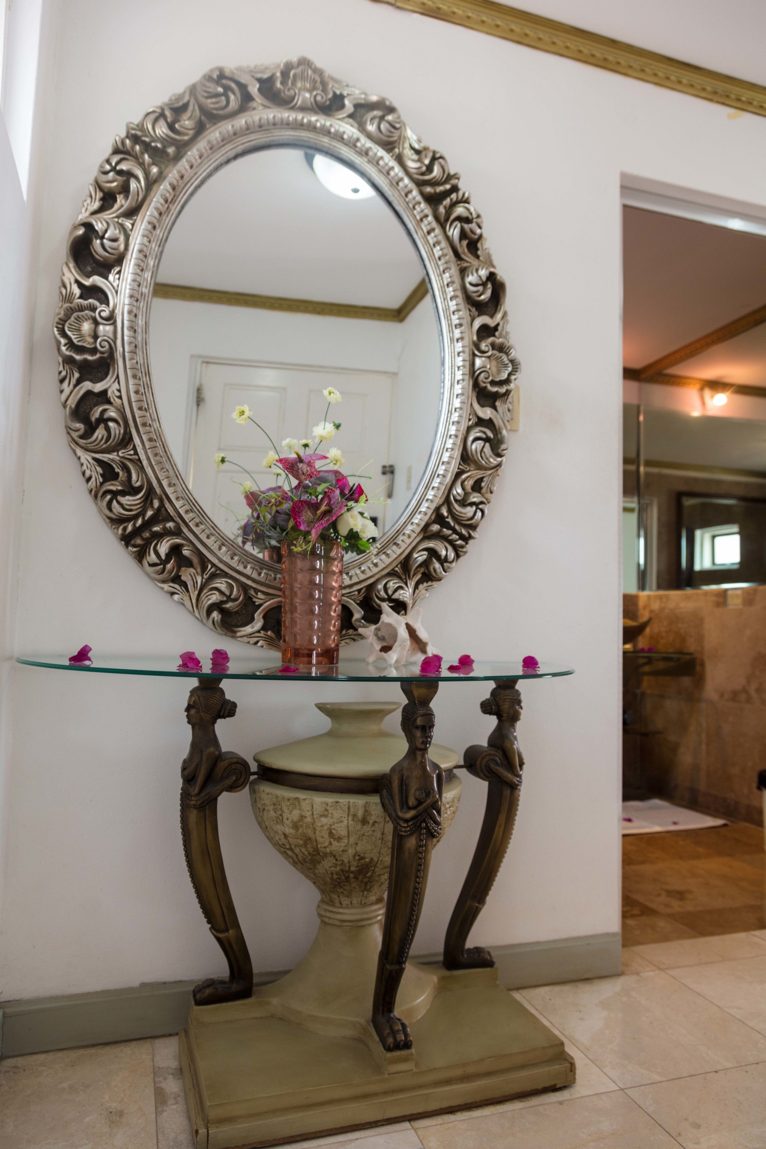 A mirror with an ornate frame, a glass table with a vase of flowers, and a decorative base.