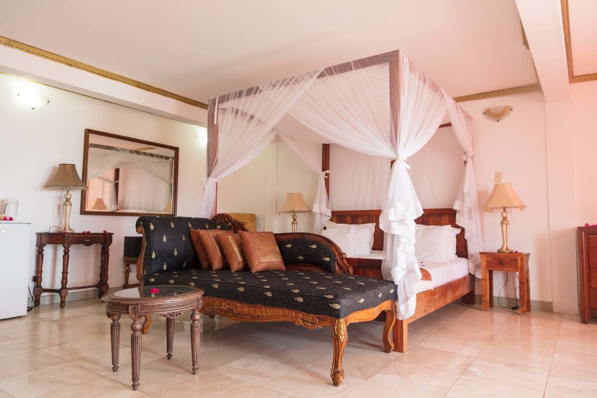 A four-poster canopy bed with a dark patterned sofa and pillows, a small round table, and two bedside lamps.
