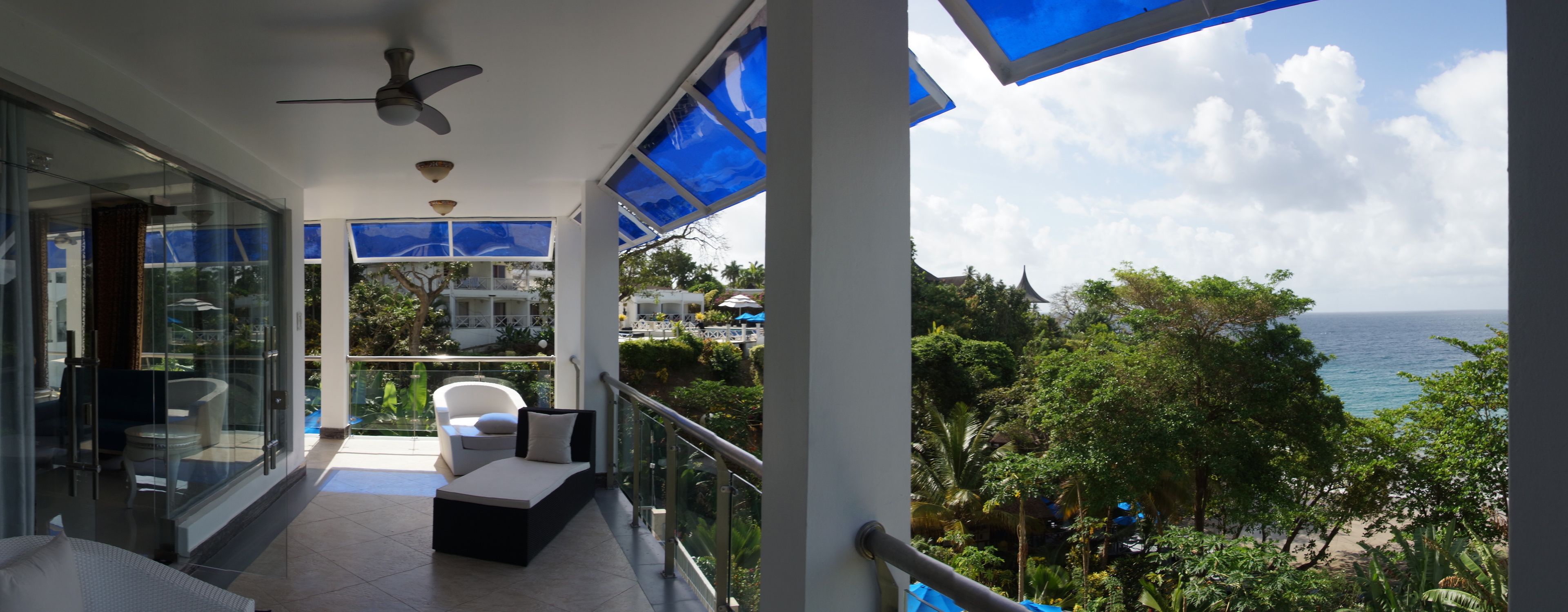A balcony with a glass railing, a ceiling fan, and a view of the ocean and greenery.