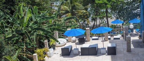 A poolside area with lounge chairs and umbrellas, surrounded by tropical vegetation and overlooking a body of water.