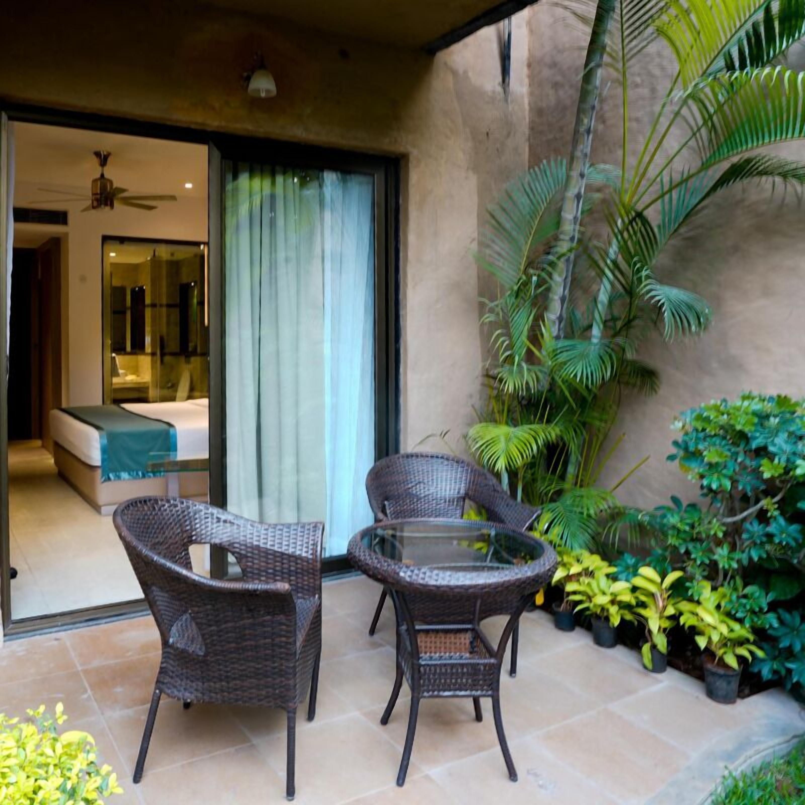 A patio area with wicker chairs and a glass table, a bedroom with a bed and a bathroom visible through a glass door, and various potted plants.