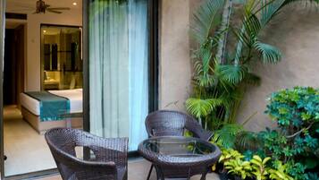 A patio area with wicker chairs and a glass table, a bedroom with a bed and a bathroom visible through a glass door, and various potted plants.