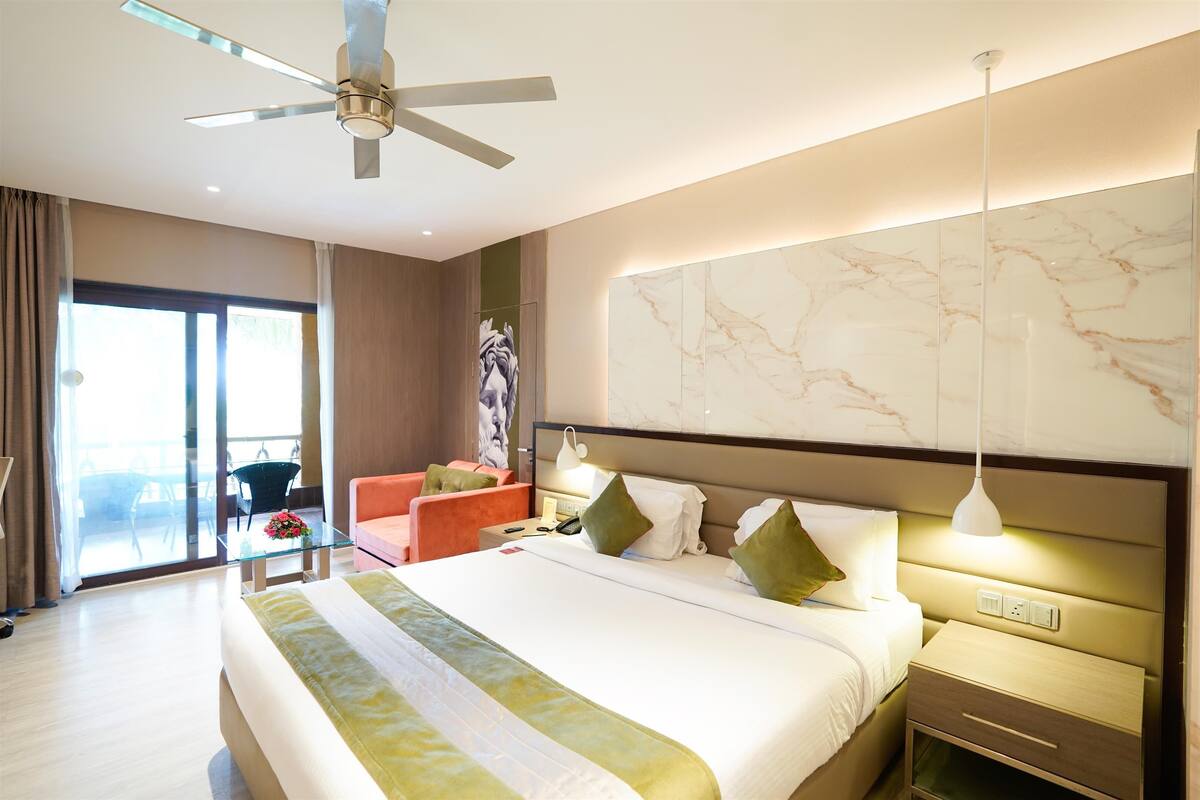 Un hotel moderno con una cama grande, una mesa de vidrio con un florero, un ventilador de techo y vistas a través de la puerta de cristal.
