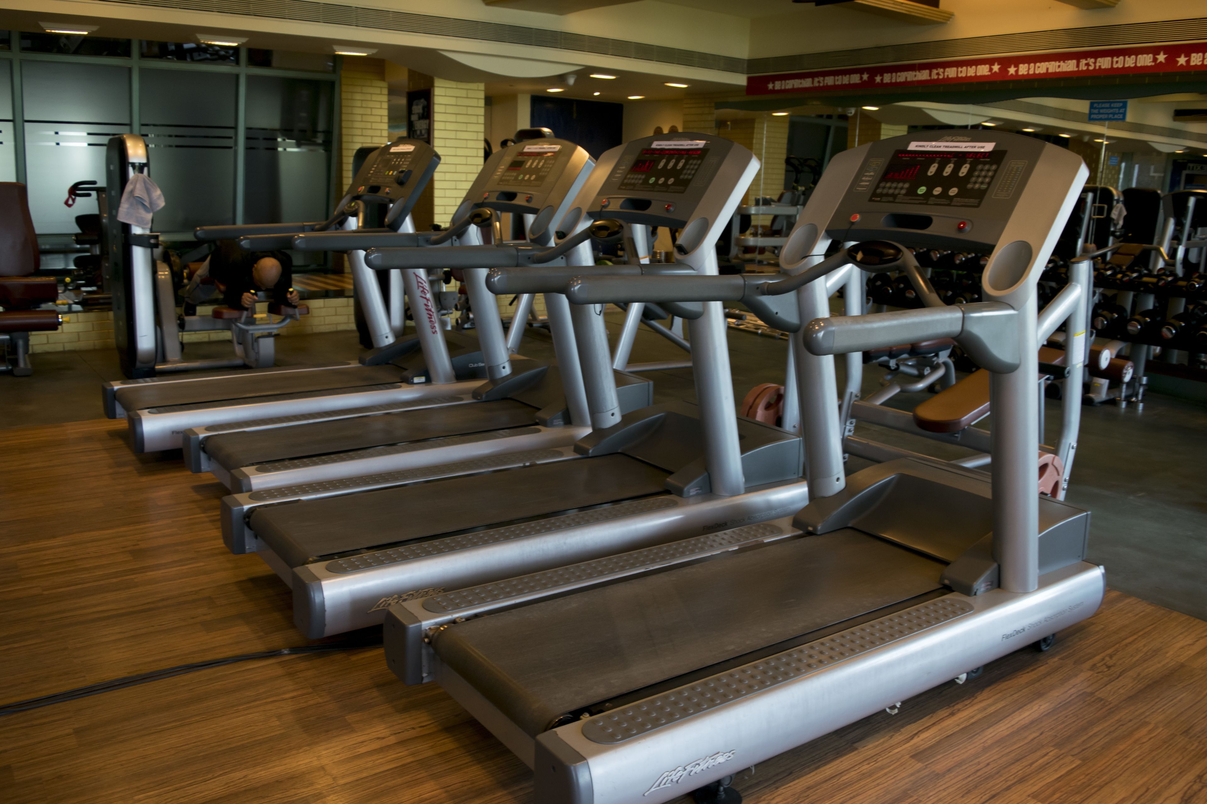 A row of treadmills in a gym with a wooden floor and various exercise equipment in the background.