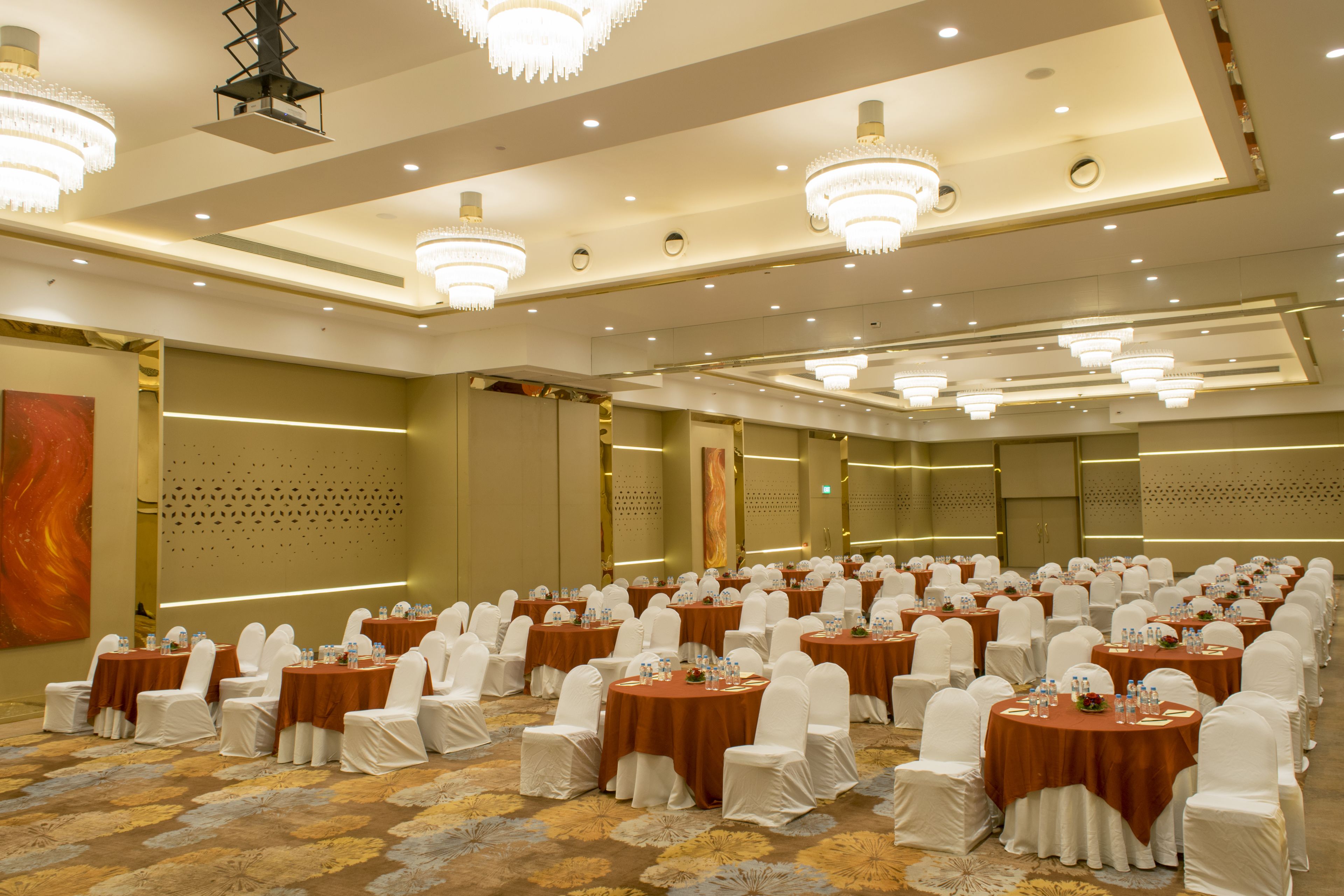 A banquet hall with round tables covered in brown tablecloths, white chairs, and chandeliers.