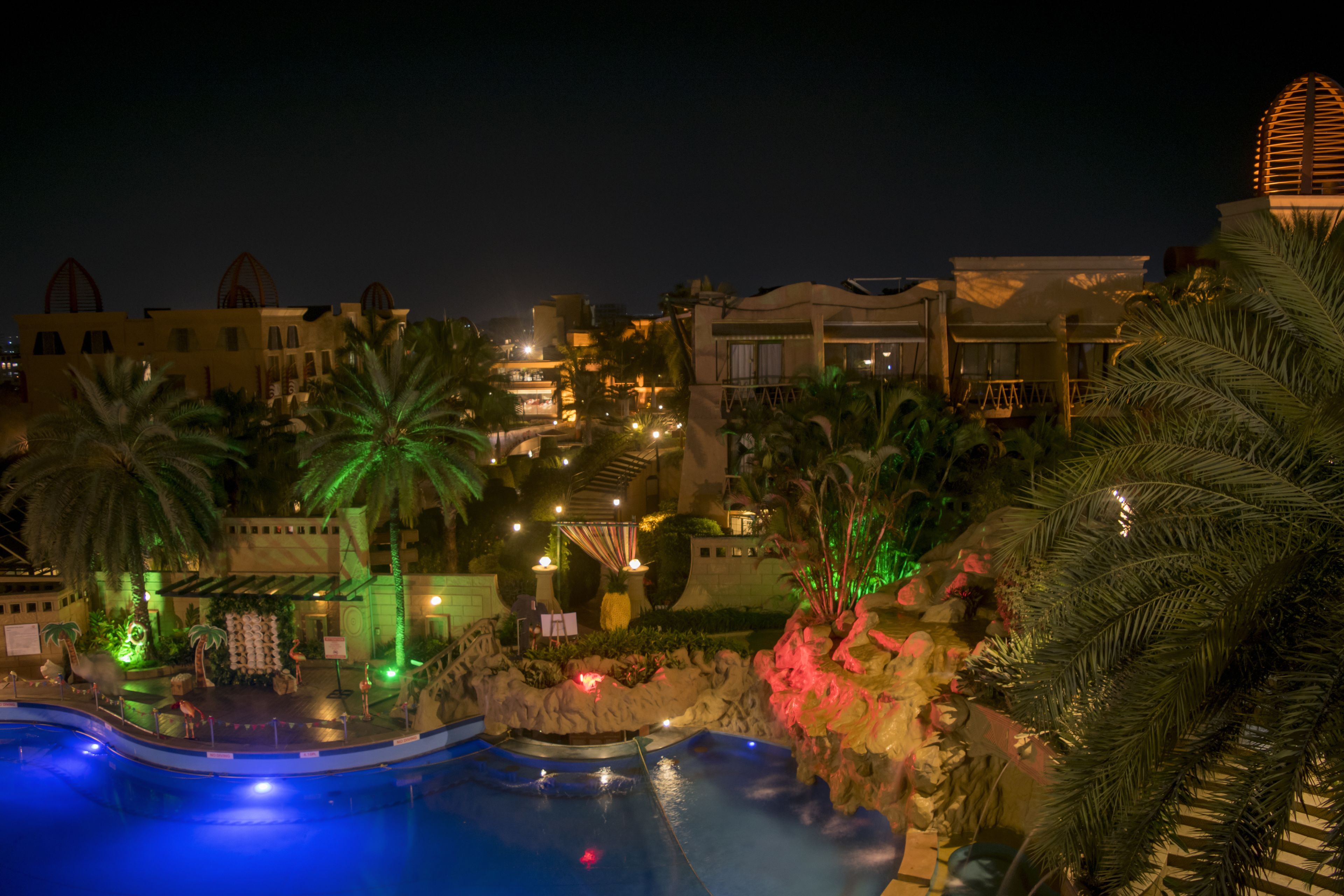 Area piscina di un resort di notte, con palme, un palco e edifici illuminati.
