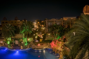 Zona de piscina de un complejo turístico de noche, con palmeras, un escenario y edificios iluminados.