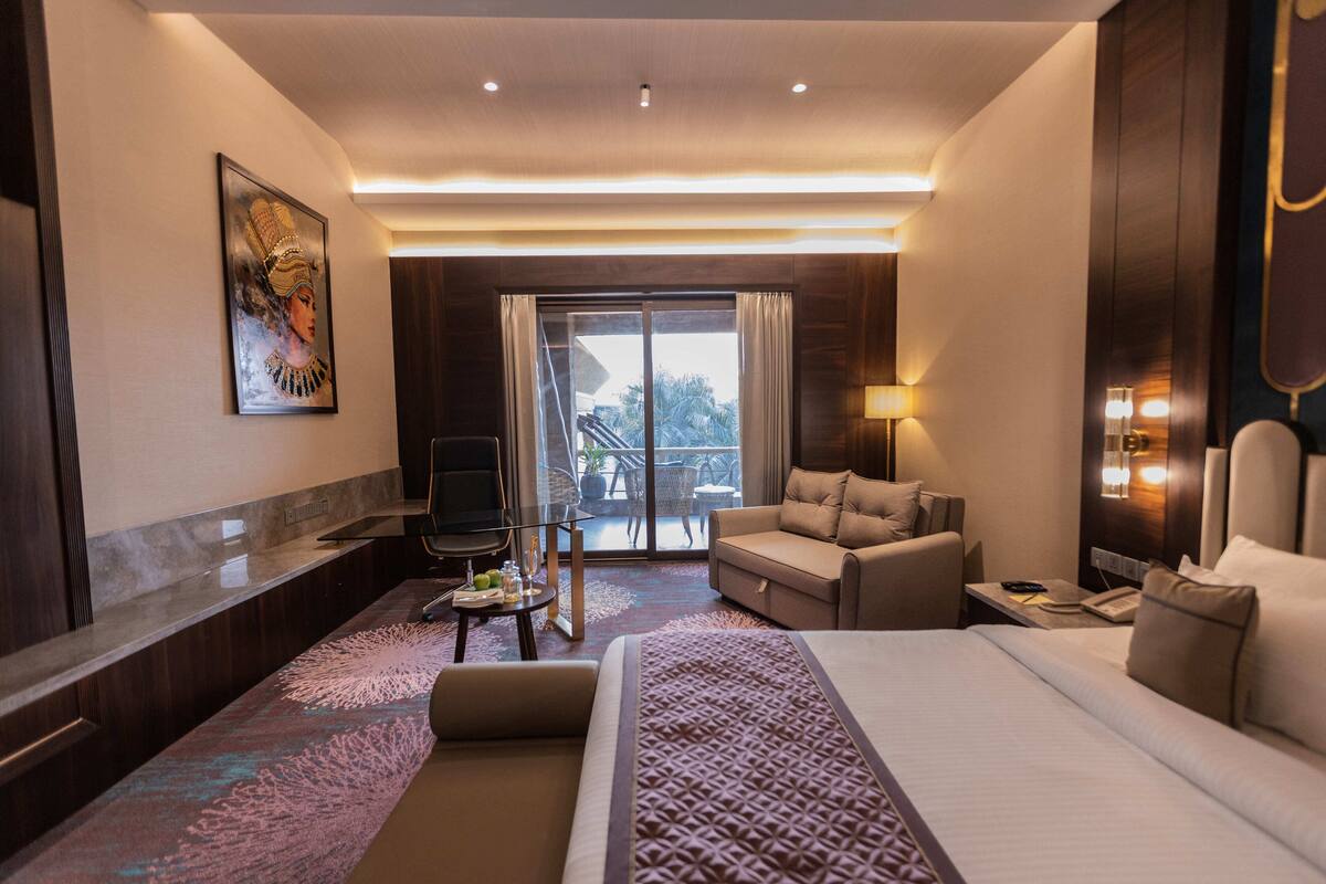 Habitación de hotel con una cama grande, un sofá, un escritorio, un televisor y un balcón con vista a palmeras.