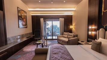 A hotel room with a large bed, a sofa, a desk, a TV, and a balcony with a view of palm trees.
