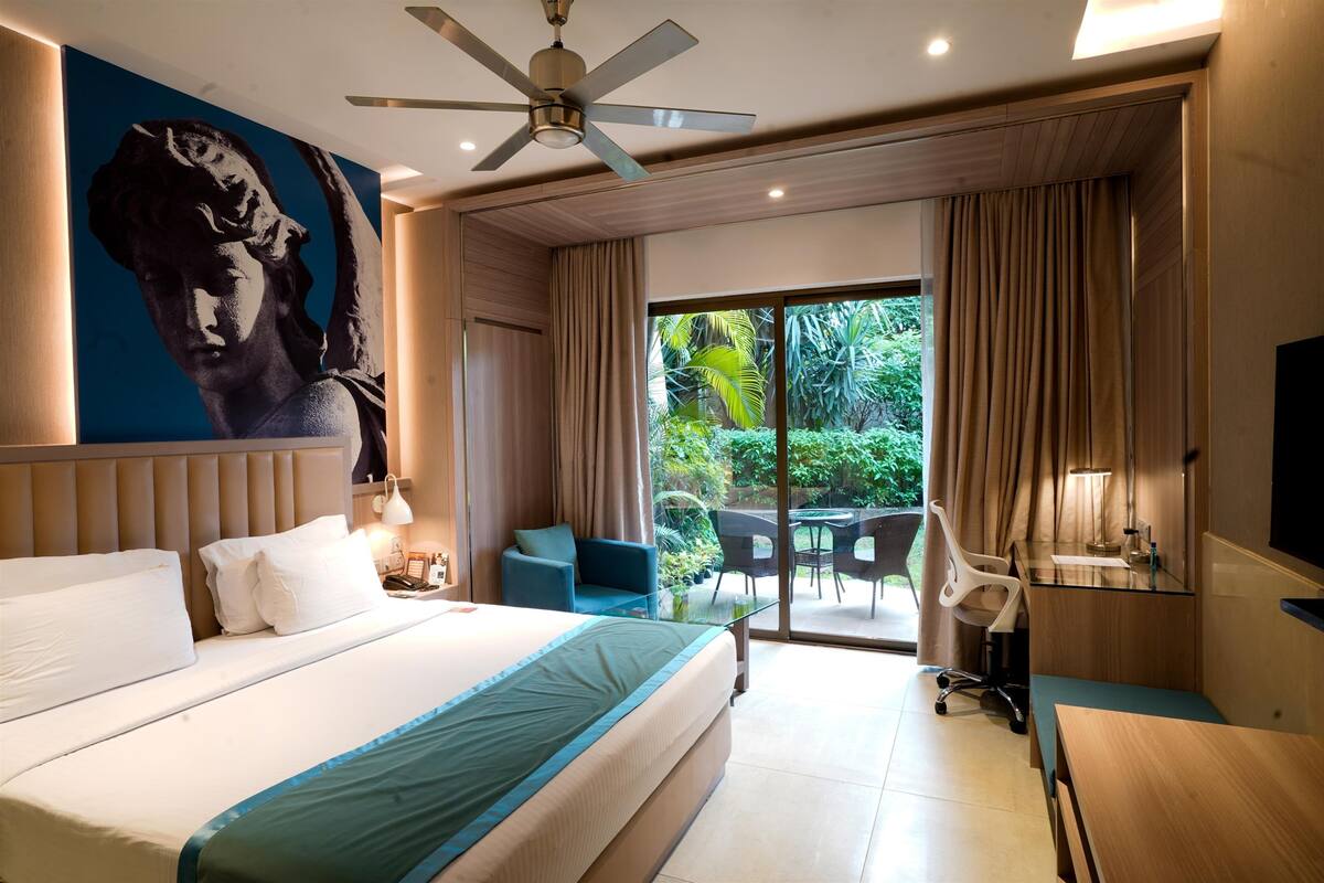 Un cuarto de hotel con una cama grande, un ventilador de techo, un cuadro, un escritorio, una silla y vistas al exterior.