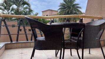 A balcony with wicker furniture and a rope barrier, overlooking palm trees and buildings.