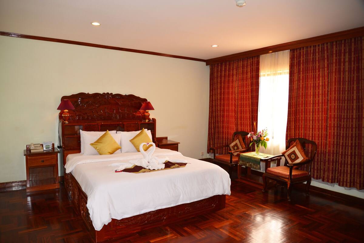 Una cama grande con cabecera de madera, una mesita de noche, una silla y una pequeña mesa con un jarrón de flores.