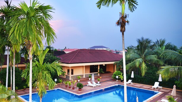A pool area with lounge chairs, palm trees, and a building with a red roof. - Angkor Hotel (Siem Reap)