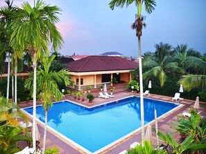 A pool area with lounge chairs, palm trees, and a building with a red roof. - Angkor Hotel (Siem Reap)
