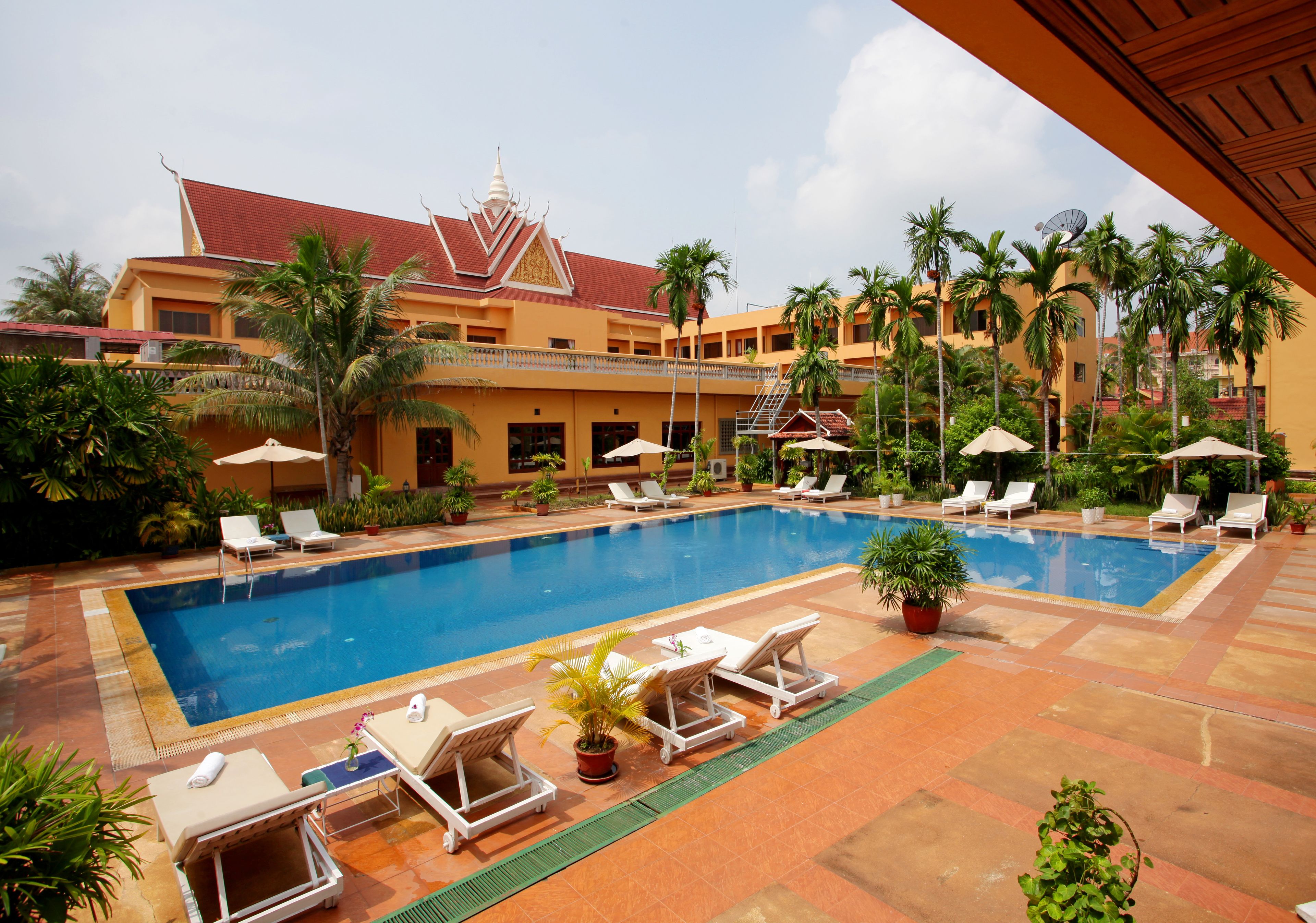 A swimming pool with lounge chairs and umbrellas, surrounded by palm trees and a building with a red roof.