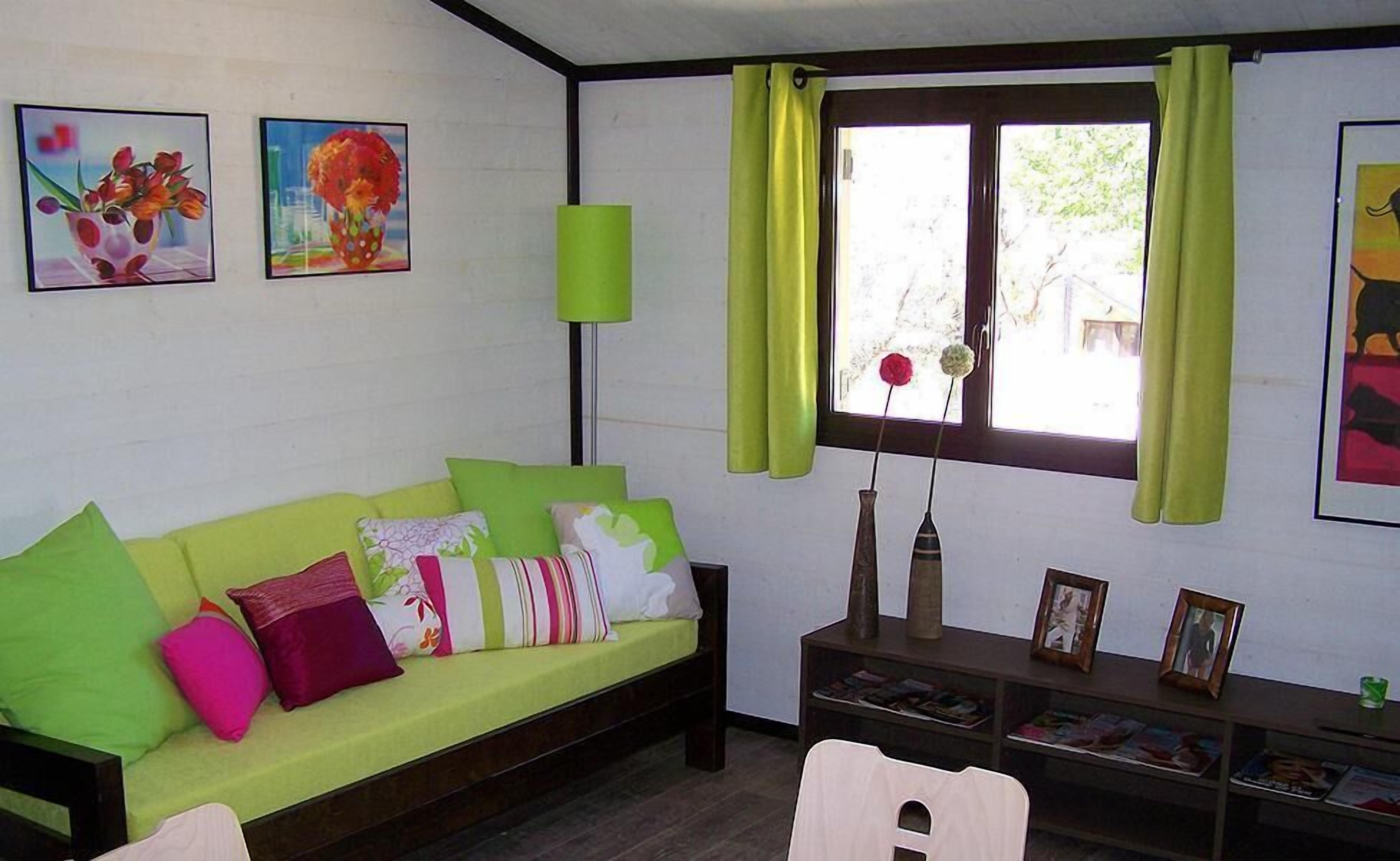 Una habitación con un sofá verde, una mesita con un jarrón y un marco de fotos, y una ventana con cortinas.