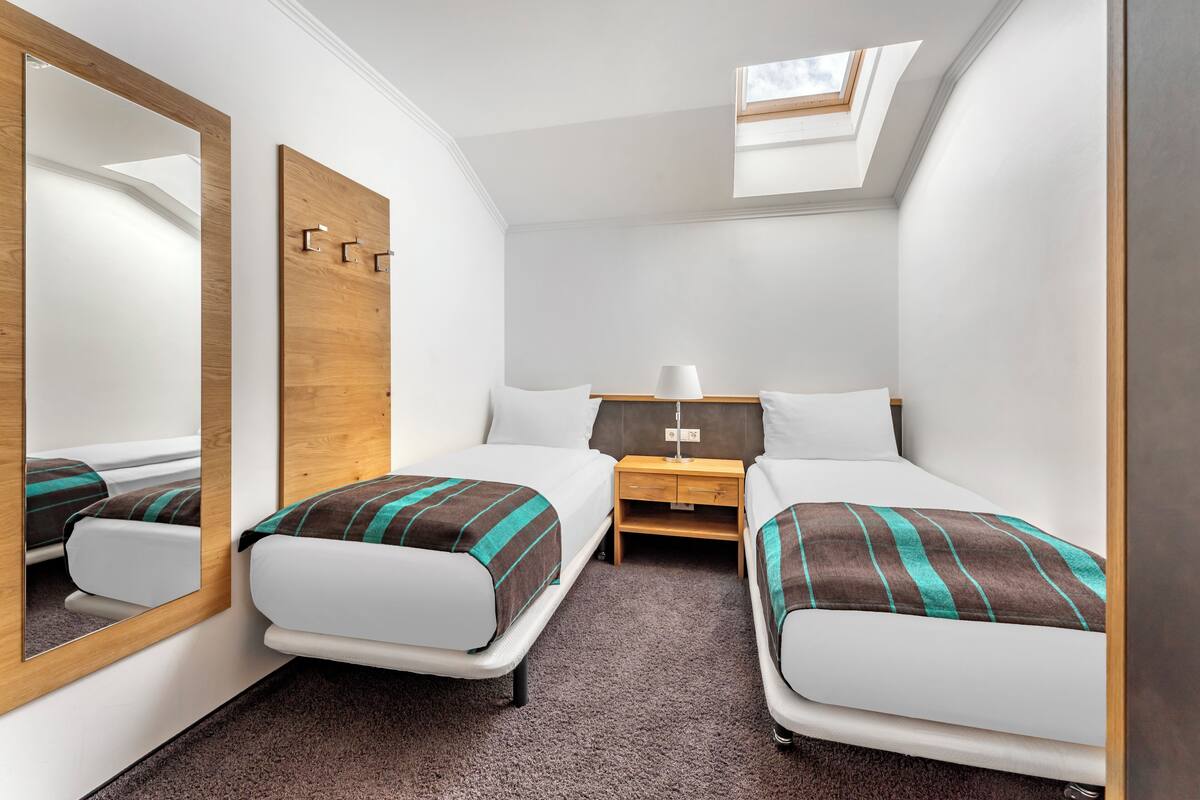 A compact hotel room with two single beds, a wooden headboard, a nightstand with a lamp, and a skylight.