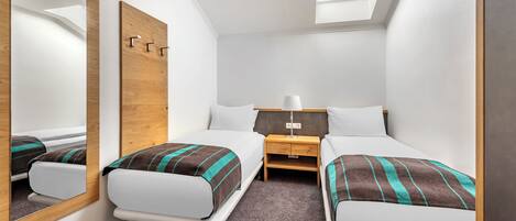 A compact hotel room with two single beds, a wooden headboard, a nightstand with a lamp, and a skylight.