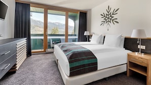 A hotel room with a bed, a TV, a desk, and a view of mountains through a sliding door. - Alpine Club (Schladming)