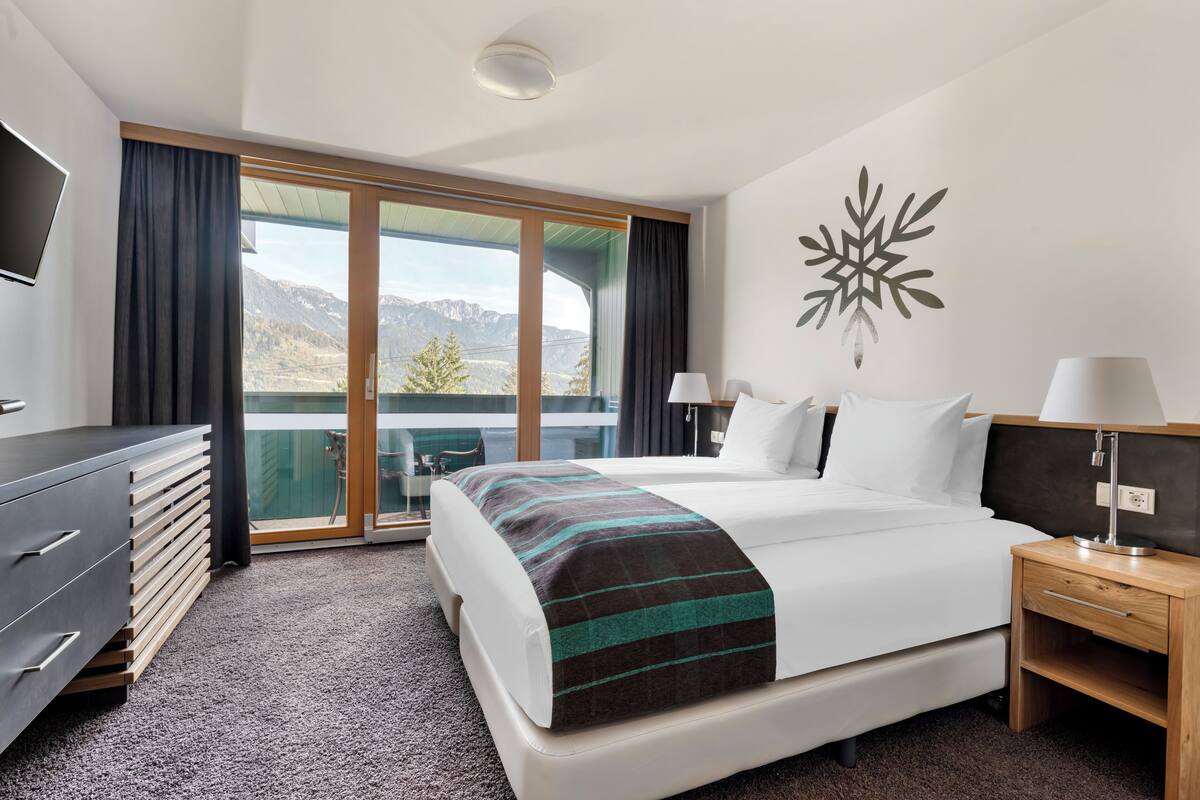 A hotel room with a bed, a TV, a desk, and a view of mountains through a sliding door.