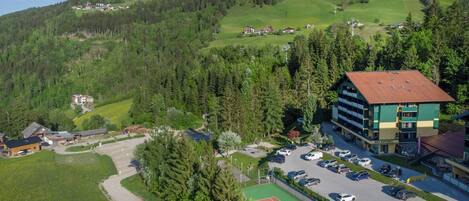 A hotel with a tennis court, surrounded by greenery and a mountainous backdrop.