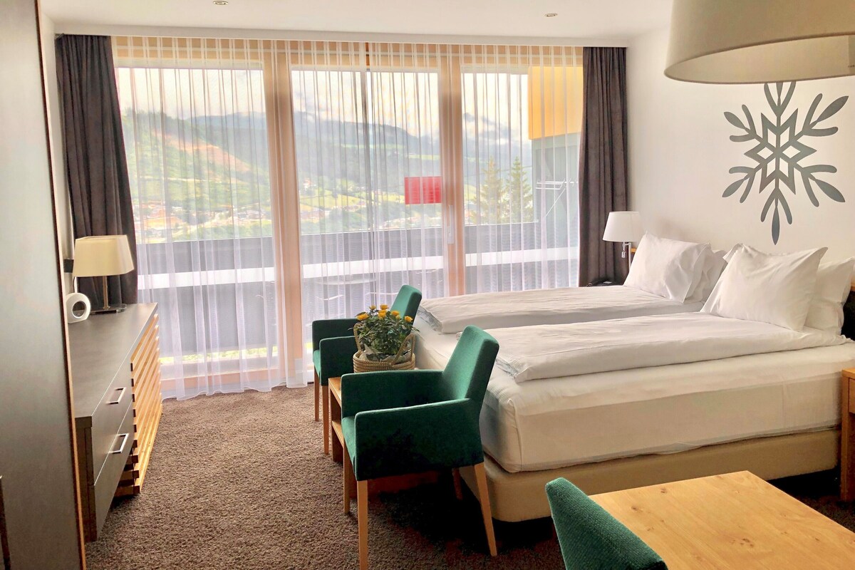 A hotel room with a bed, two chairs, a desk, a lamp, and a view of mountains through large windows.