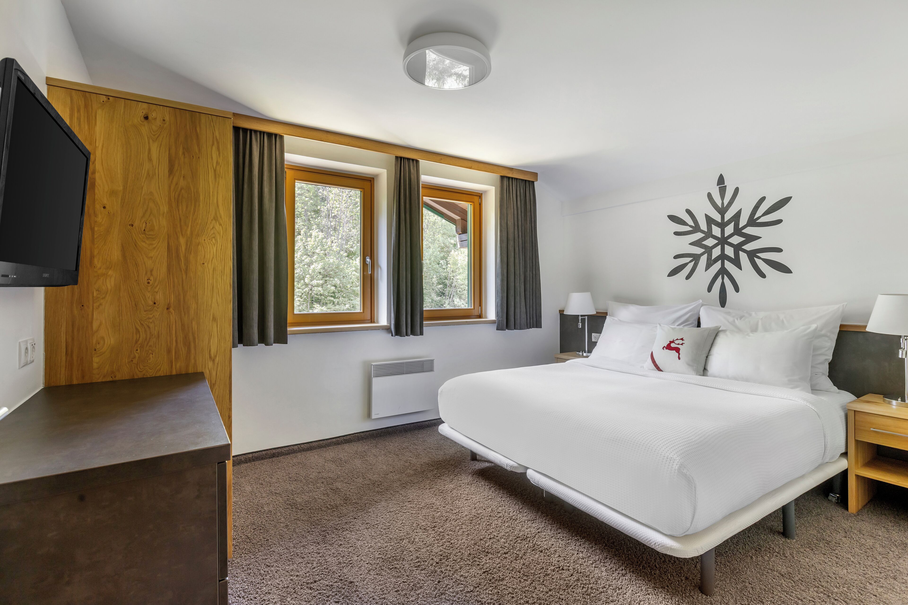 Una habitación de hotel con una cama grande, televisión, pared de paneles de madera, ventana con cortinas, un adorno de pared y una mesita de noche.