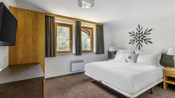Una habitación de hotel con una cama grande, televisión, pared de paneles de madera, ventana con cortinas, un adorno de pared y una mesita de noche.