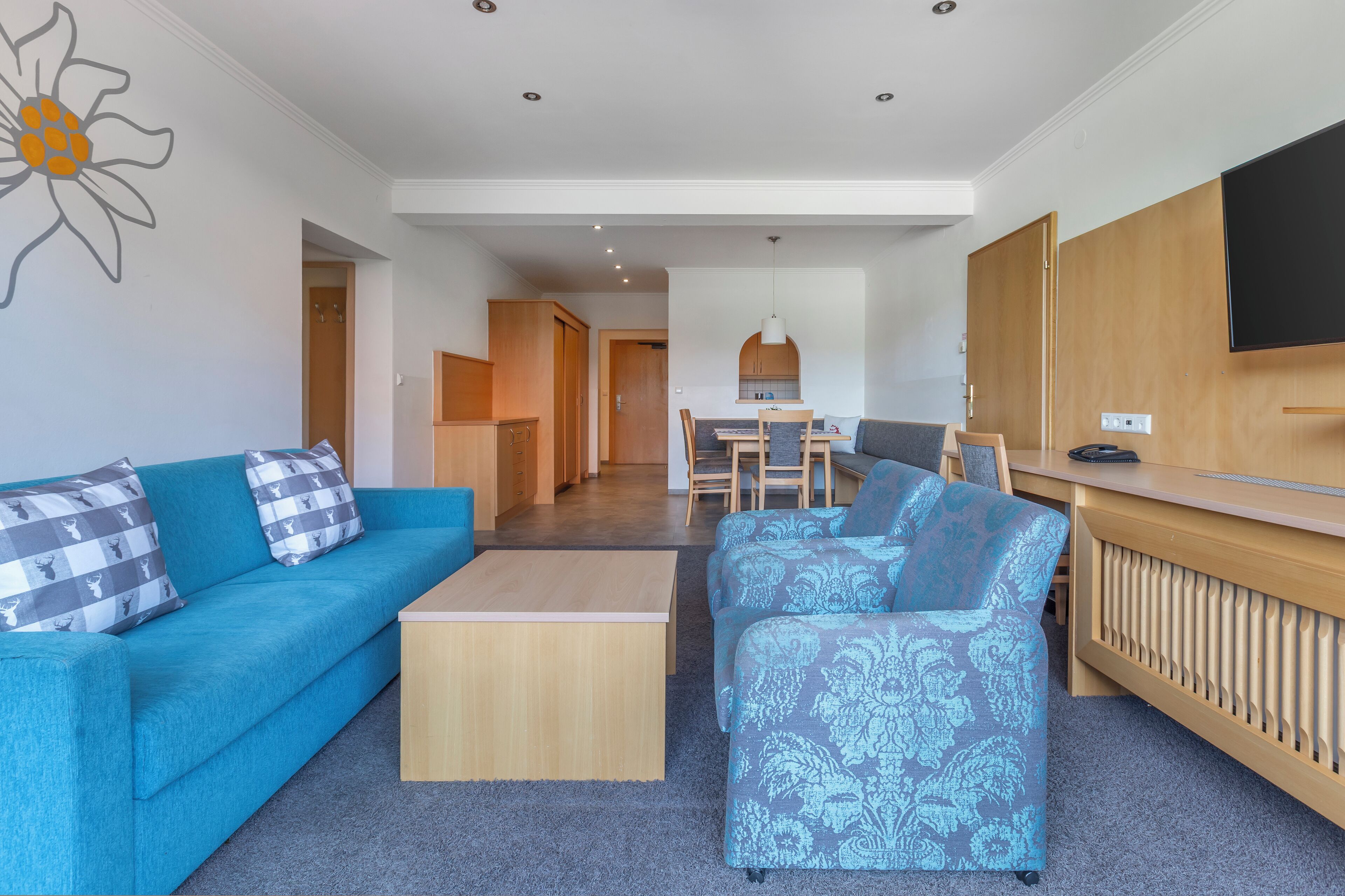 Una sala moderna con un sofá azul, un sillón estampado y una mesa de centro de madera. Se observa un comedor con una mesa y sillas, y una cocina al fondo.
