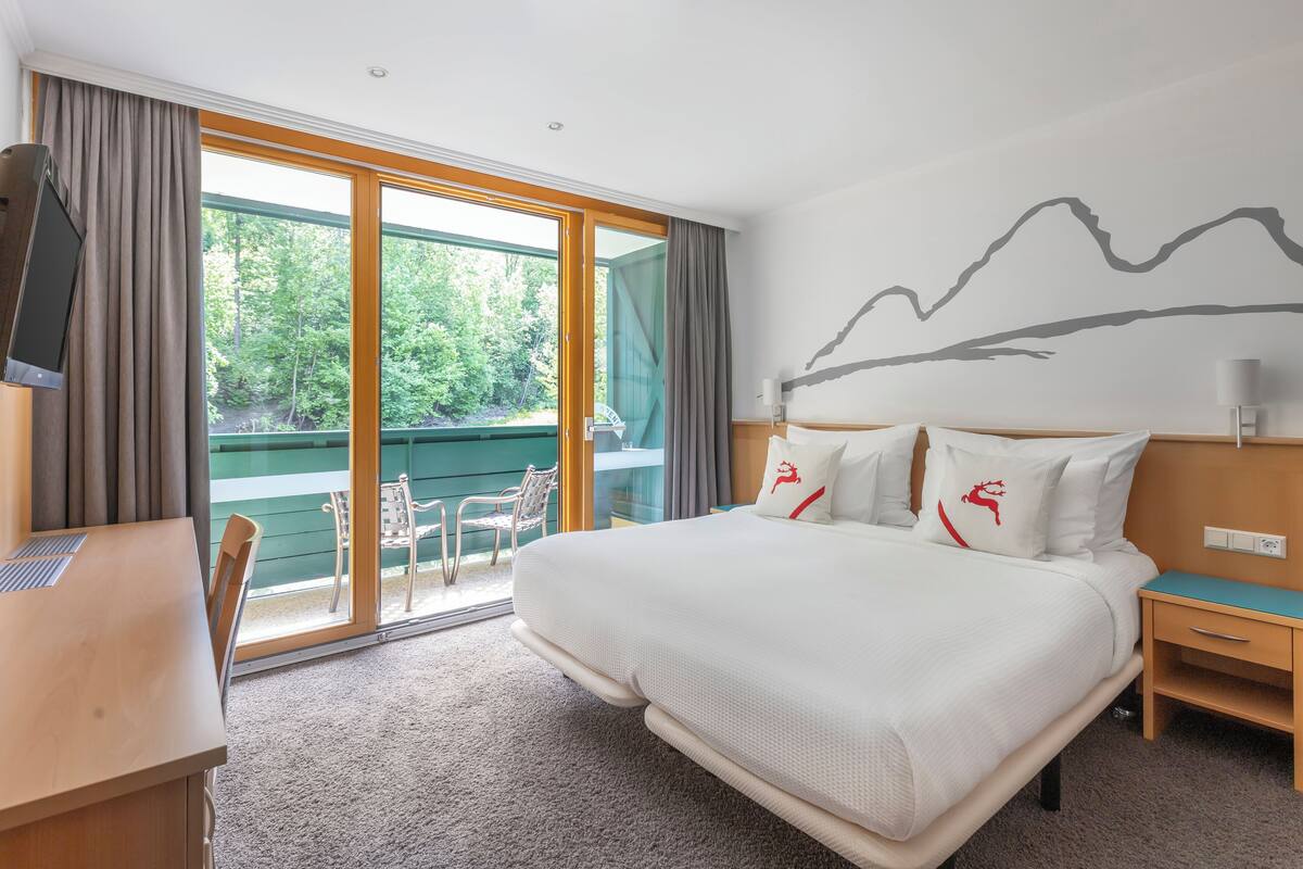A hotel room with a large bed, a desk, and a view of greenery through the sliding glass door.