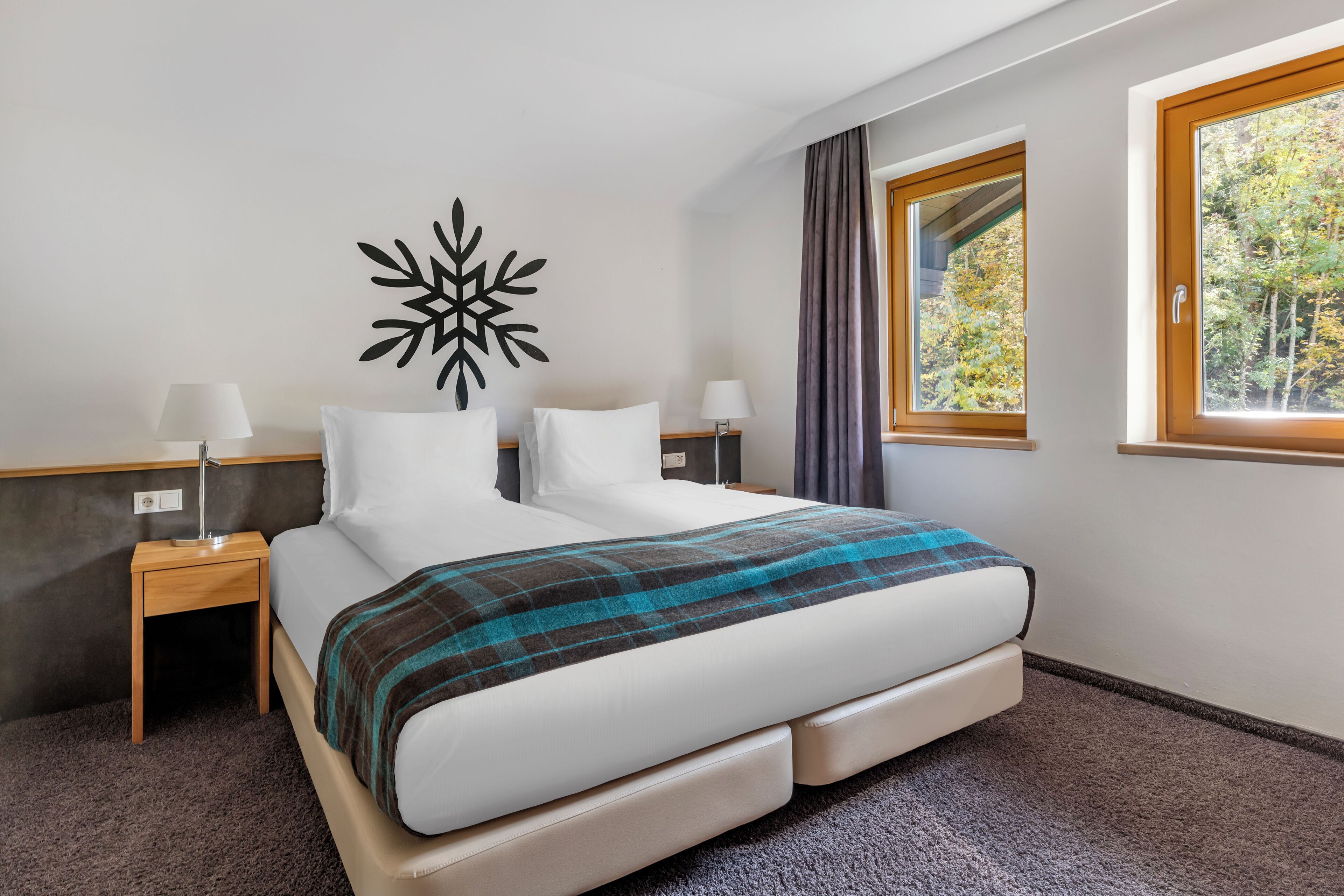 Habitación de hotel con cama, dos mesitas de noche, un adorno en la pared, ventana con cortinas y vistas a árboles.