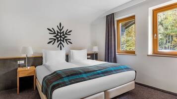 Habitación de hotel con cama, dos mesitas de noche, un adorno en la pared, ventana con cortinas y vistas a árboles.