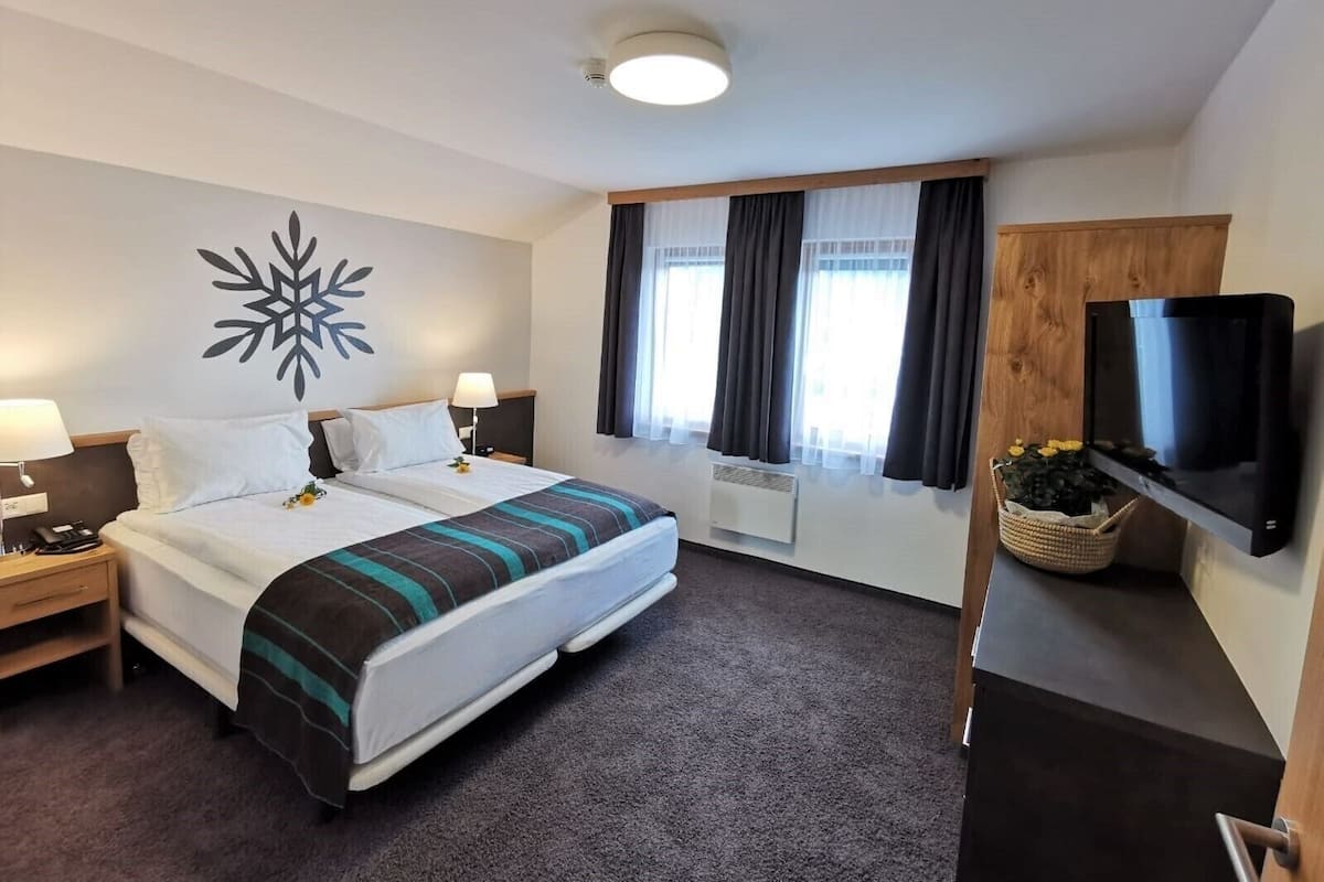 A hotel room with a bed, two bedside tables, a flat-screen TV, a window with curtains, and a decorative wall piece.