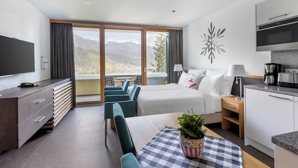 A modern hotel room with a bed, a dining table with chairs, a kitchen area, and a balcony with a mountain view. - Alpine Club (Schladming)