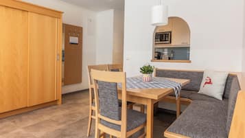 Una habitación compacta con una mesa de comedor de madera, dos sillas, un sofá, una pequeña cocina con microondas y un armario de madera.