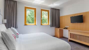 A modern hotel room with a large bed, a desk, a TV, and two windows with curtains.