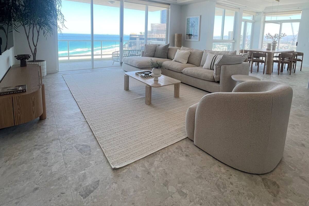 A spacious living room with a large window overlooking the ocean, a modern sofa, a coffee table, and a dining area with a view.