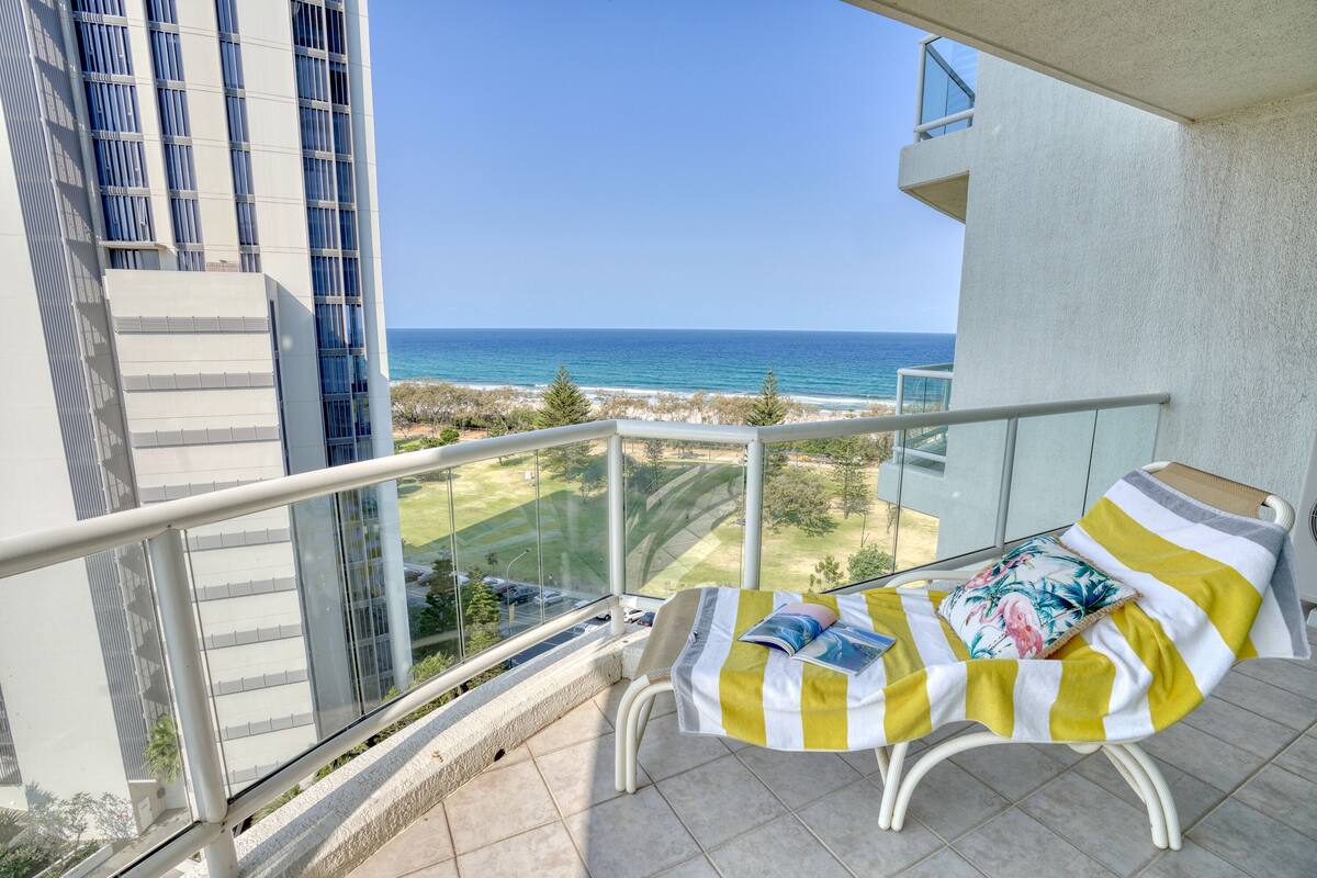 A balcony with a view of the ocean, a lounge chair with a striped blanket, and a glass railing.